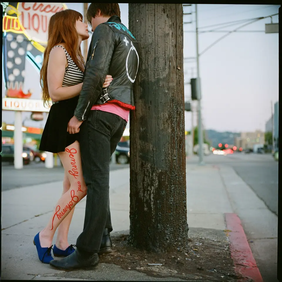 Jonathon Rice and Jenny Lewis embracing on a street against a telegraph pole. Jenny has “Jenny and Johnny” written in red down her leg..