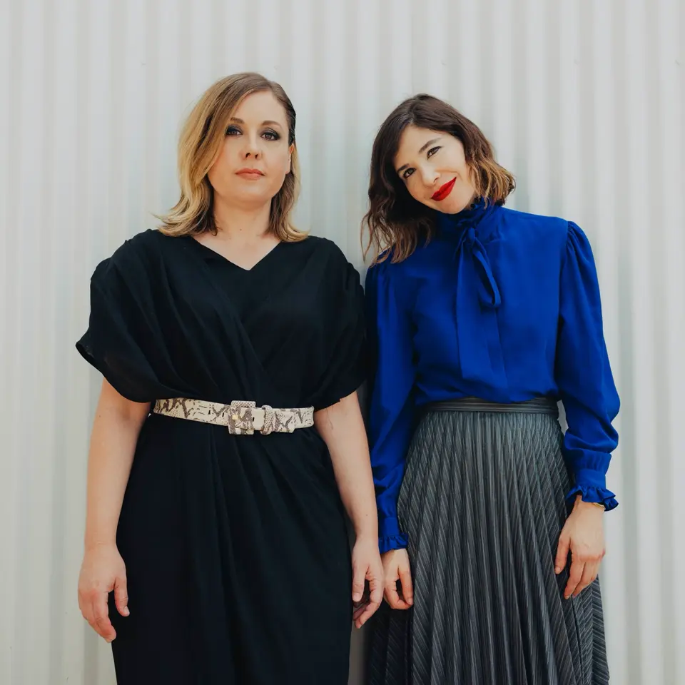 Corin Tucker right and Carrie Brownstein left, pose against a white corrugated metal sheet.