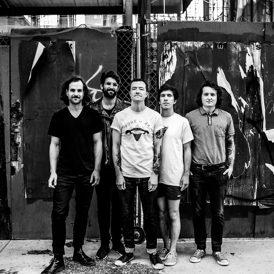 Black and white photo of the members of Touché Amore against a metal link fence.