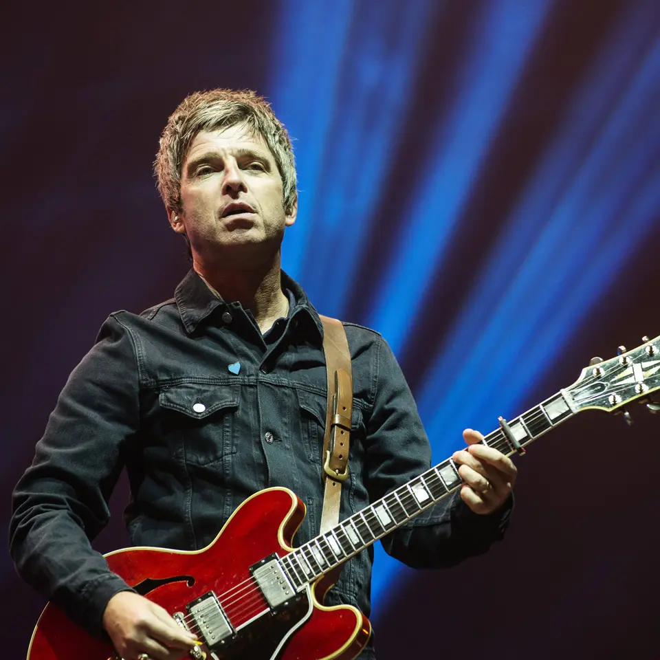 Noel Gallagher live on stage playing a red Gibson ES-355 guitar against a dark blue backdrop.