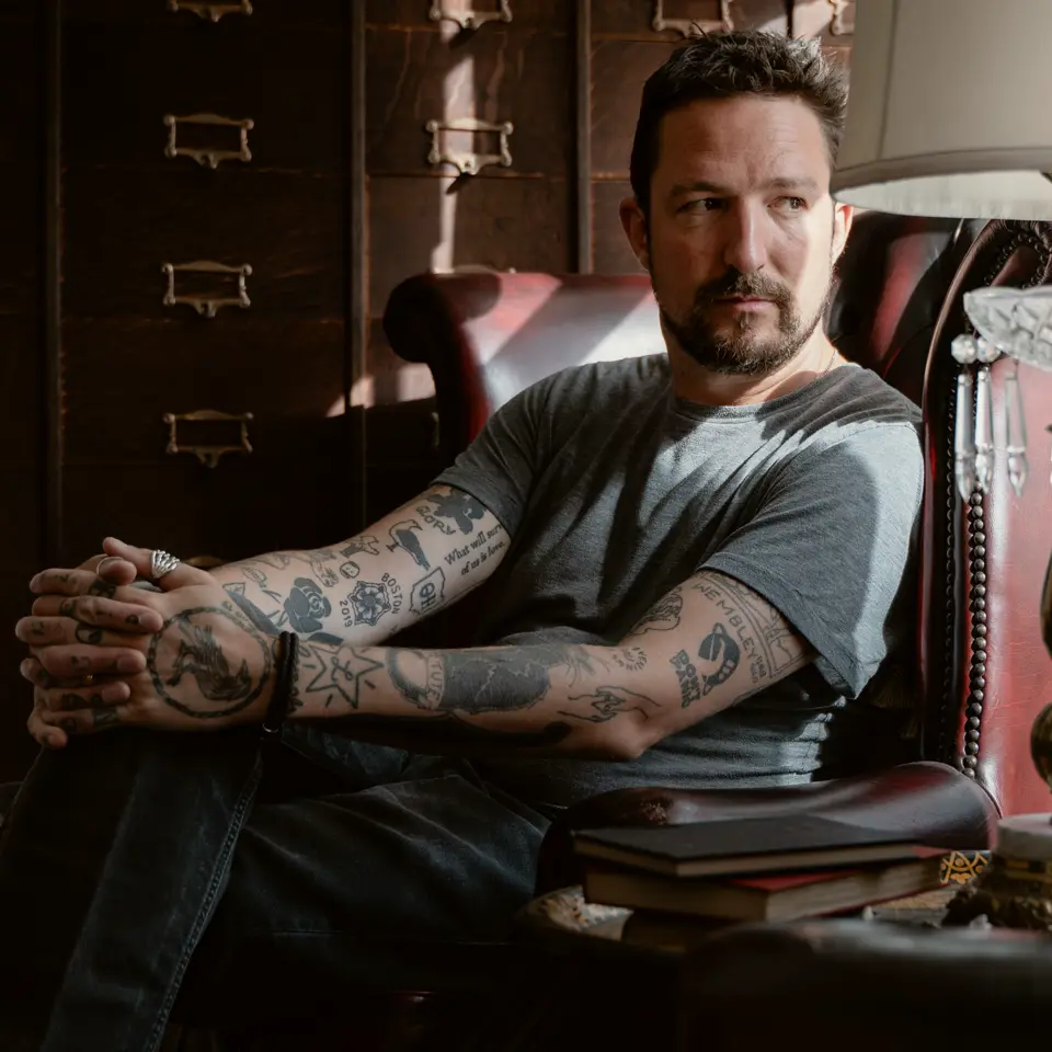 Frank Turner sat in an old room looking out the window with a lamp and books on the side table.